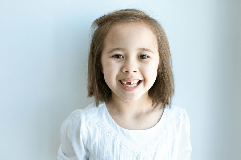 pexels-photo-6182764-6182764 Cheerful young girl showing a missing tooth, wearing a white top against a plain background.